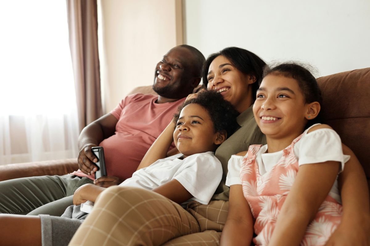 Família (casal e duas crianças) sentada no sofá assistindo televisão